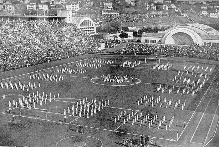 Fotografia histórica em preto e branco do Pacaembu com o campo tomado por uma apresentação coletiva e arquibancadas lotadas